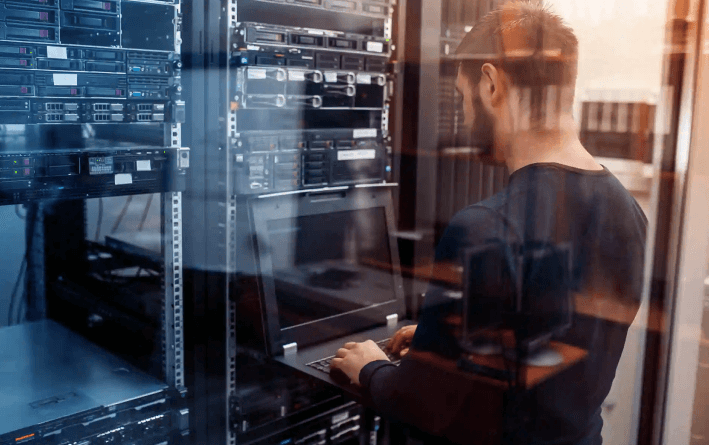 A person is standing in a server room, using a laptop connected to server racks filled with network and computer equipment—an environment showcasing expert Cabling Services. The scene is viewed through glass, creating subtle reflections.