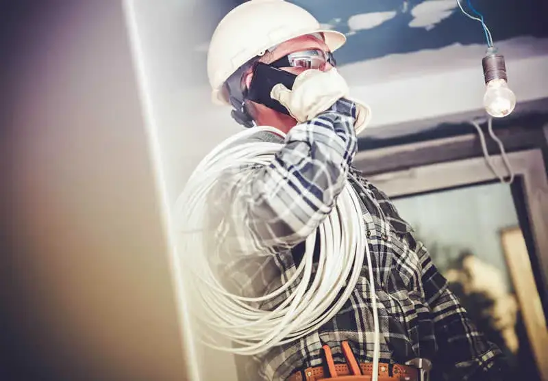 An electrician wearing a hard hat, gloves, and a plaid shirt is talking on the phone with a coil of cable over his shoulder, standing near an exposed light bulb in a room under construction—ideal for Network Cabling Services Dallas.